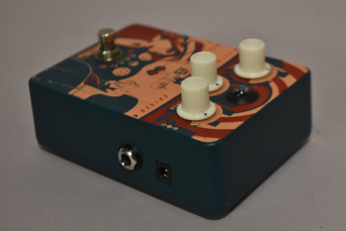 The dark teal pedal, angled from the front-left, has a beige and red graphic top, three cream knobs, a footswitch, and two input jacks.