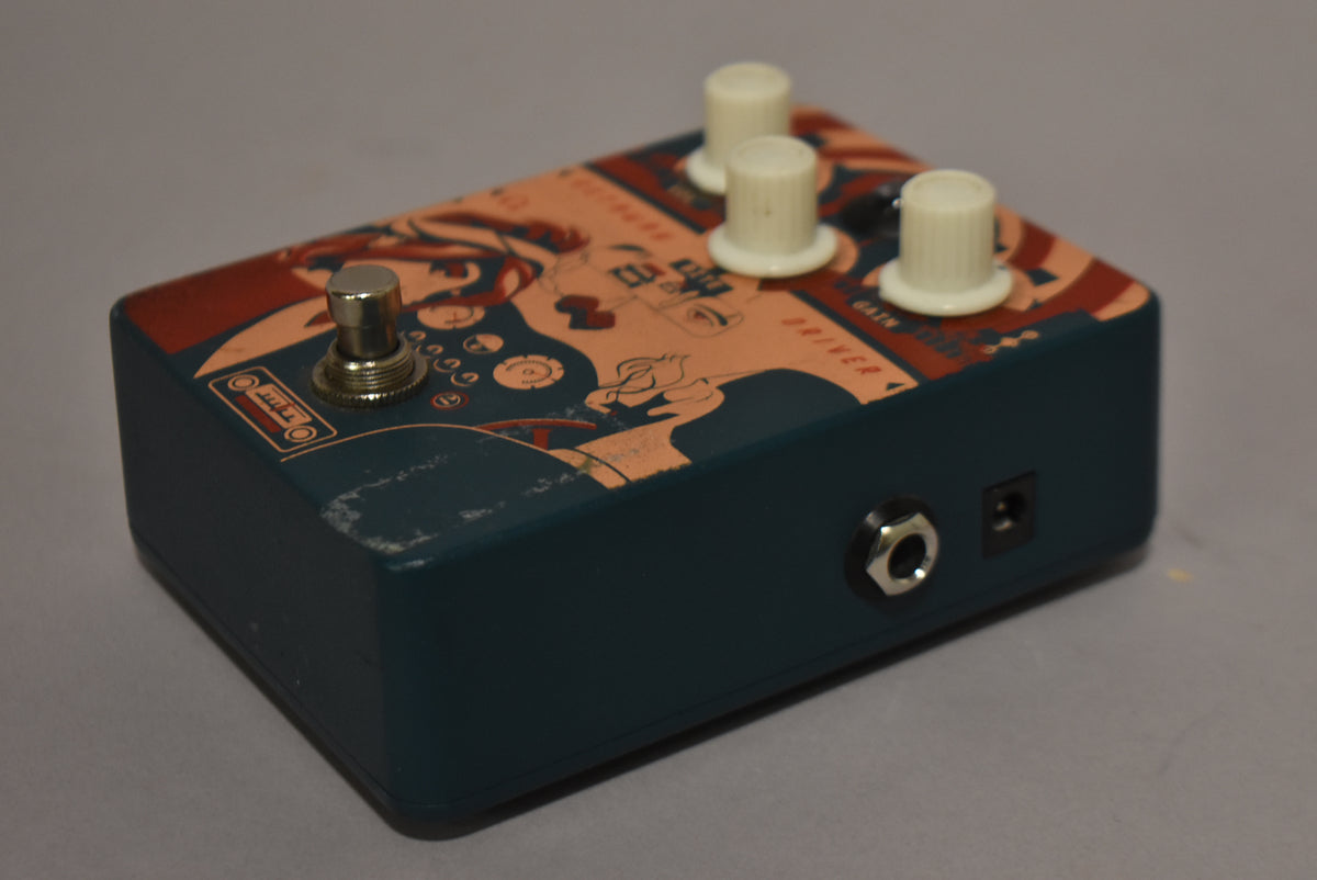 The teal pedal, angled from the right, has a peach and red graphic top, three cream knobs, a silver footswitch, and two side jacks.