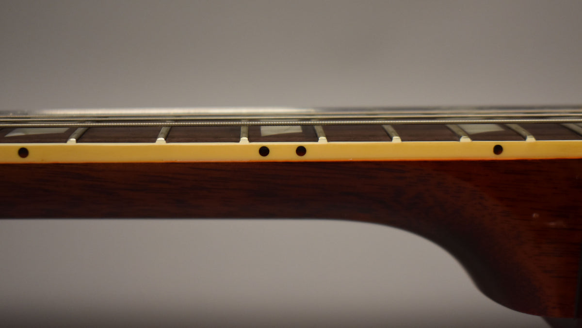 Close-up of the guitar's dark brown neck and fretboard from the side, showing cream binding with black dot markers and trapezoid inlays.