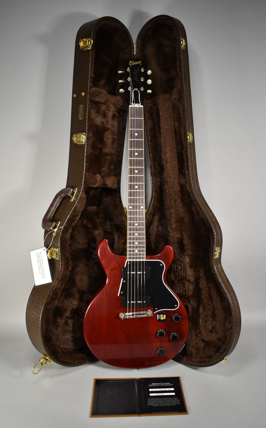 Full-length view of a cherry red double-cut guitar with a black pickguard, two P90 pickups, and four black control knobs, standing in its open brown hard case.