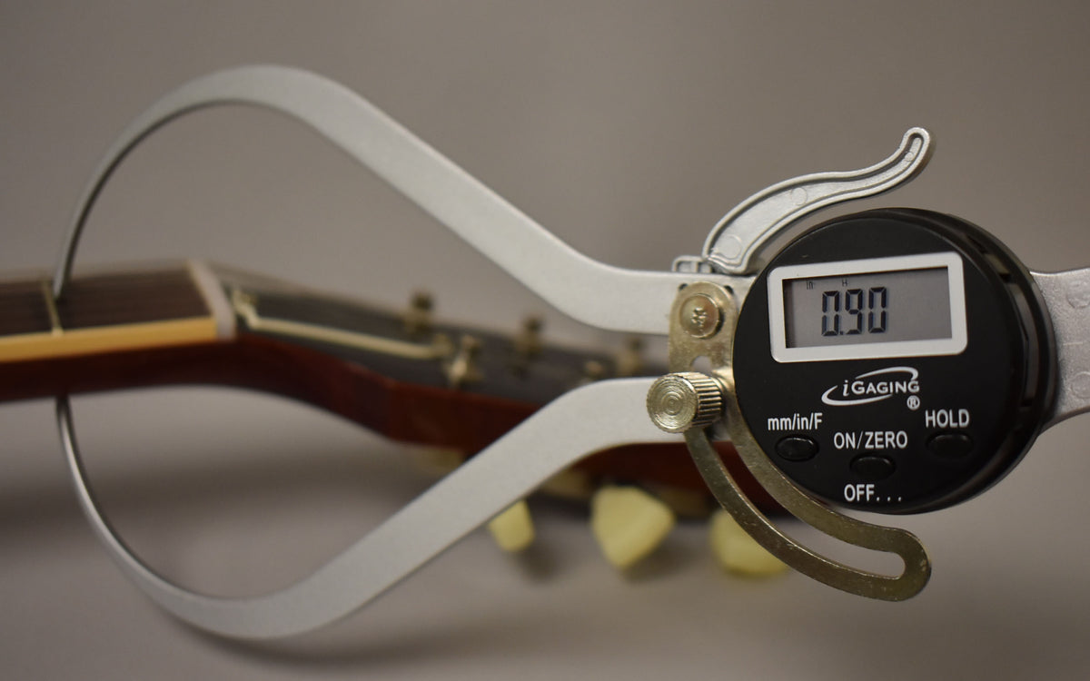 A digital caliper measures the guitar's dark brown fretboard, showing a 0.90 reading, with ivory tuning pegs blurred in the background.