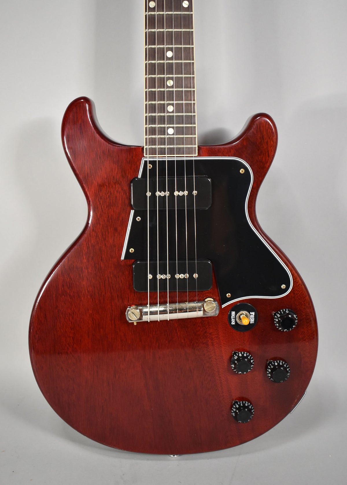 The cherry red double-cut guitar is angled slightly right, showing its dark rosewood fretboard, black pickguard, two P90 pickups, and four black control knobs.