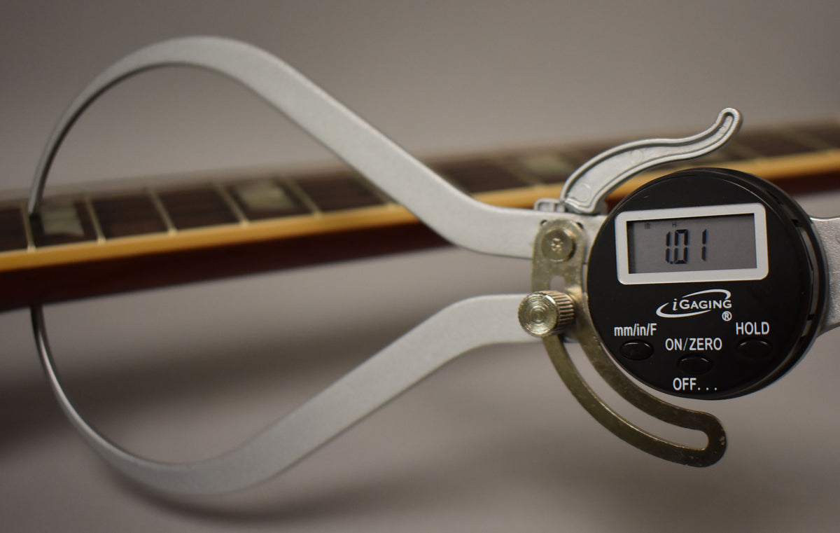A digital caliper measures the neck thickness of the guitar, showing "1.01" on its display, with the fretboard and frets visible.