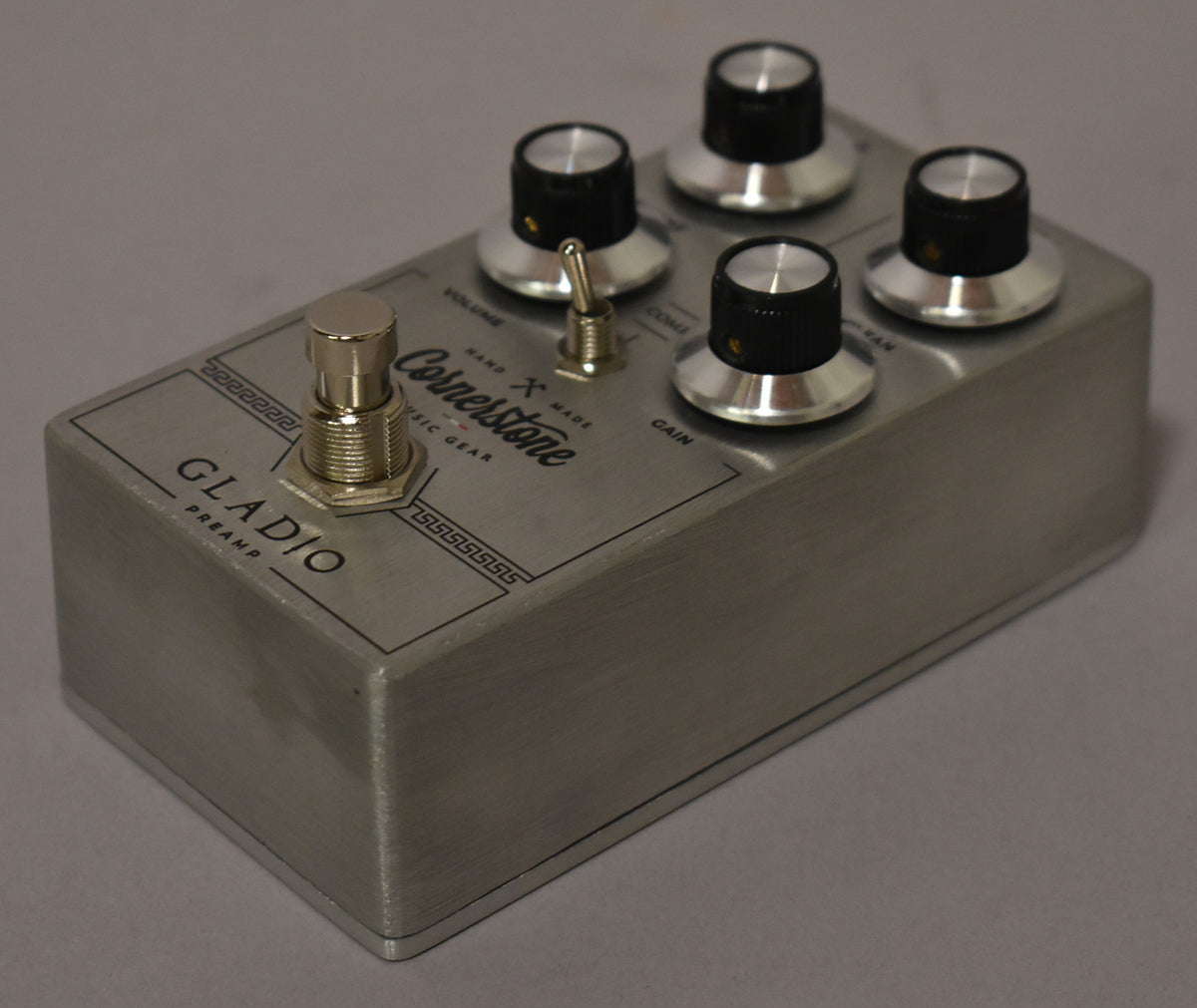 A brushed silver pedal, angled from the front right, shows a footswitch, a toggle switch, and four black and silver control knobs.