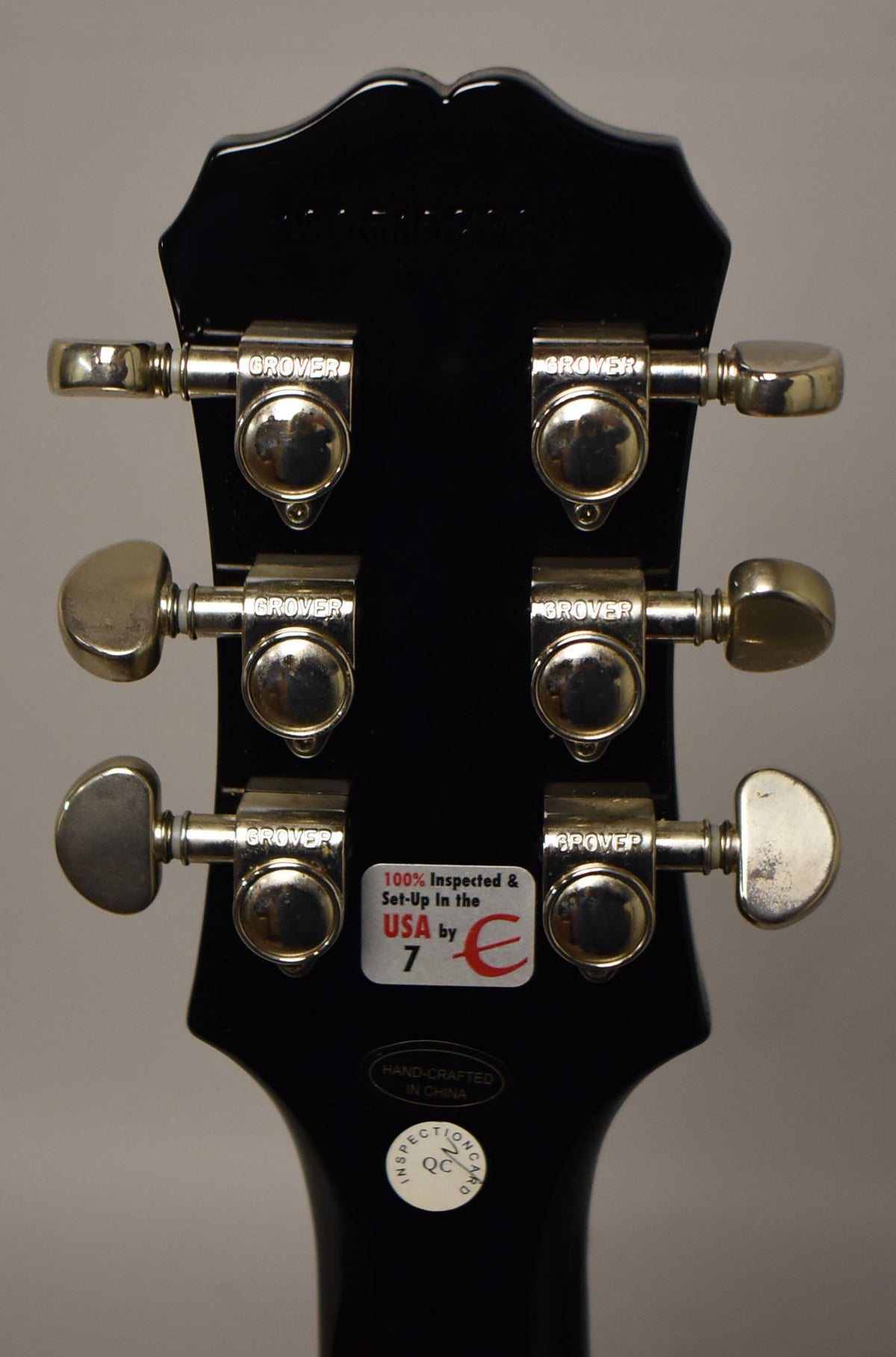 The back of a black gloss headstock with six chrome Grover tuners, showing some wear and a "100% Inspected" sticker.