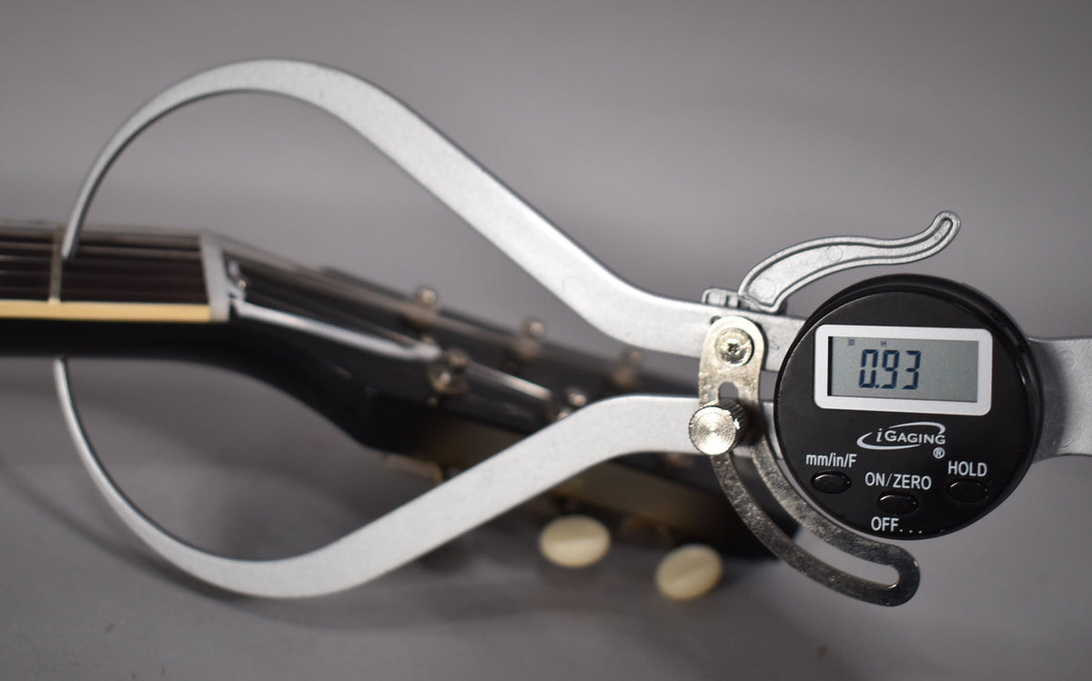 A digital caliper measures the guitar's nut, showing "0.93" on its screen, with the headstock and tuners blurred in the background.