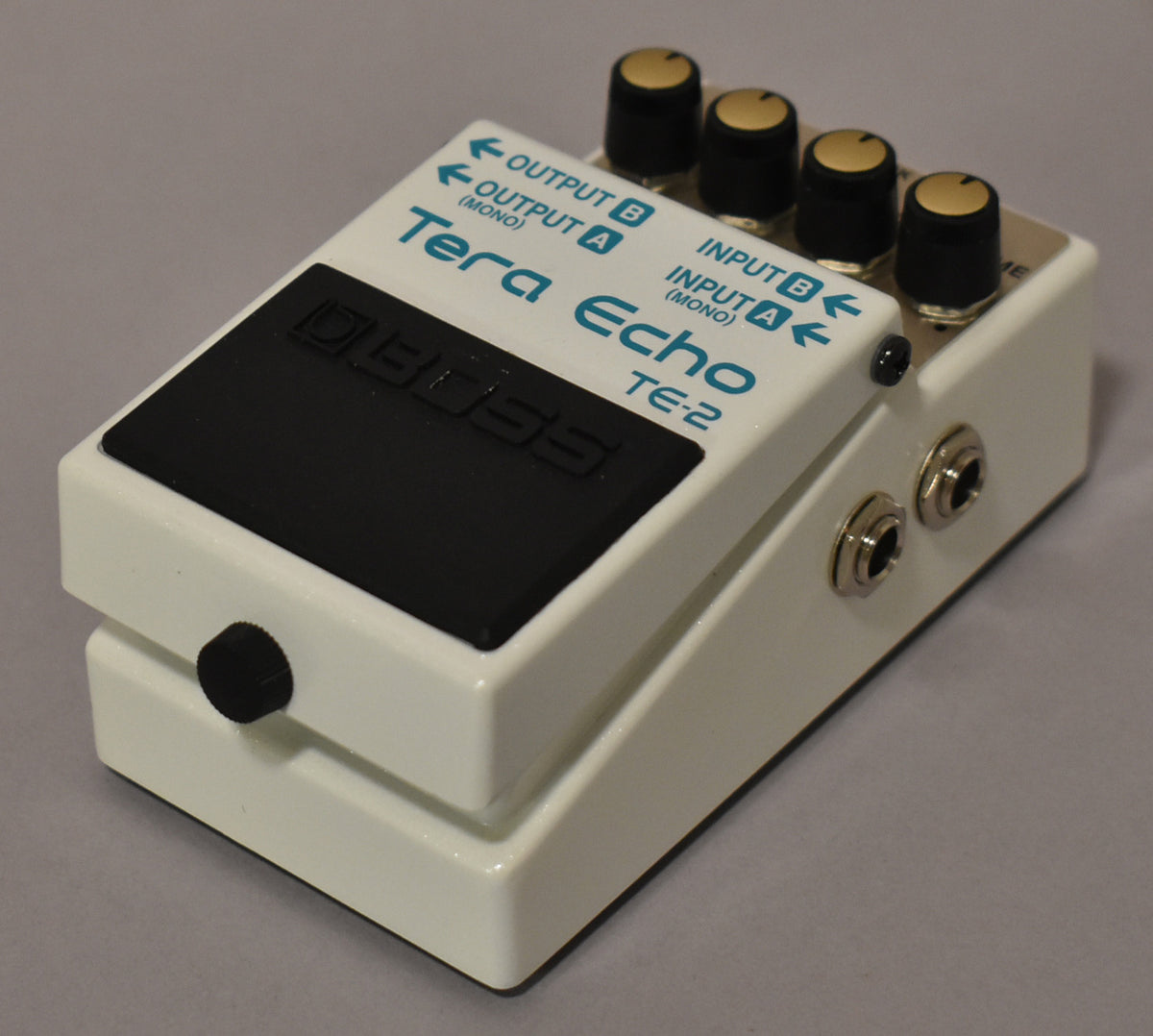 A white pedal, angled from the right, showing four black knobs with gold tops, a black footswitch, and two input jacks.