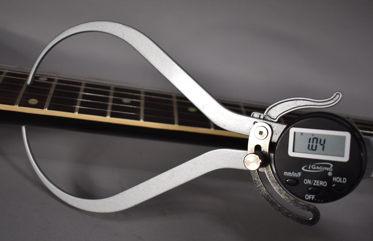 A digital caliper measures the guitar's fretboard and frets, displaying "1.04" on its screen.