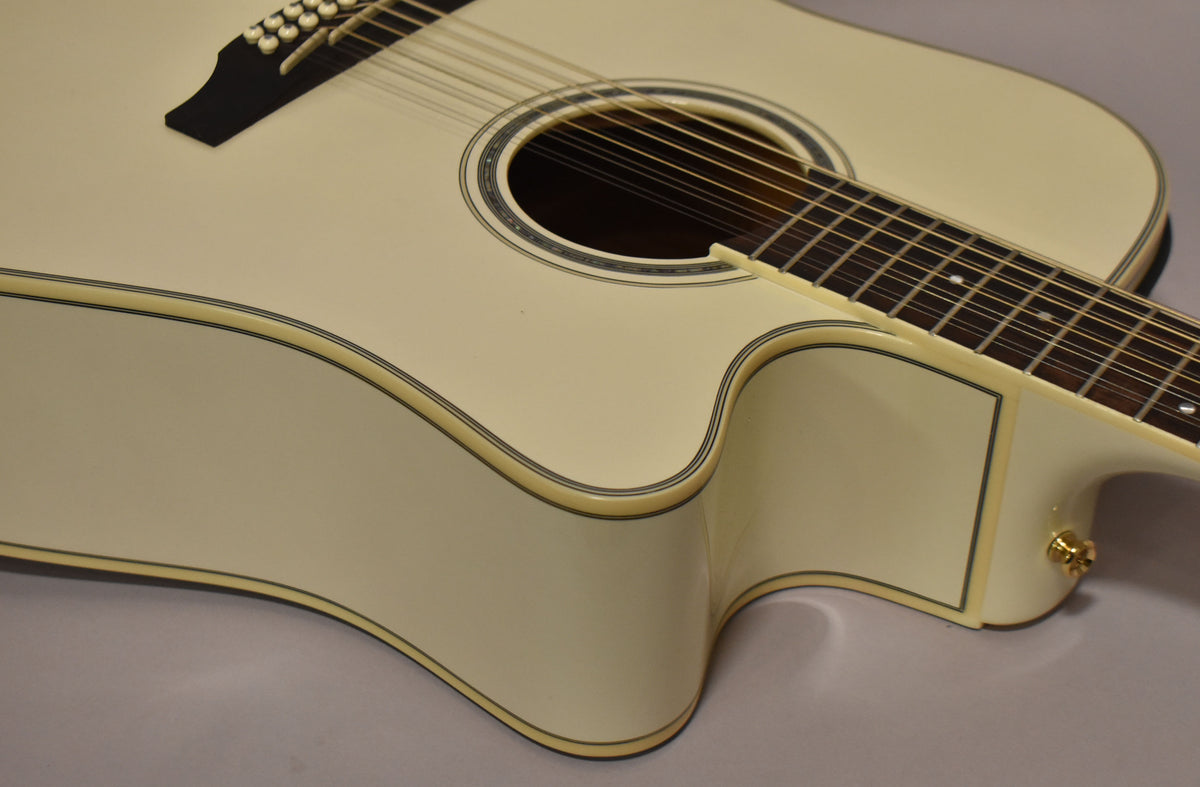 The pearl white 12-string dreadnought guitar is angled to show its cutaway, soundhole, and lower bout, with a gold strap button visible.