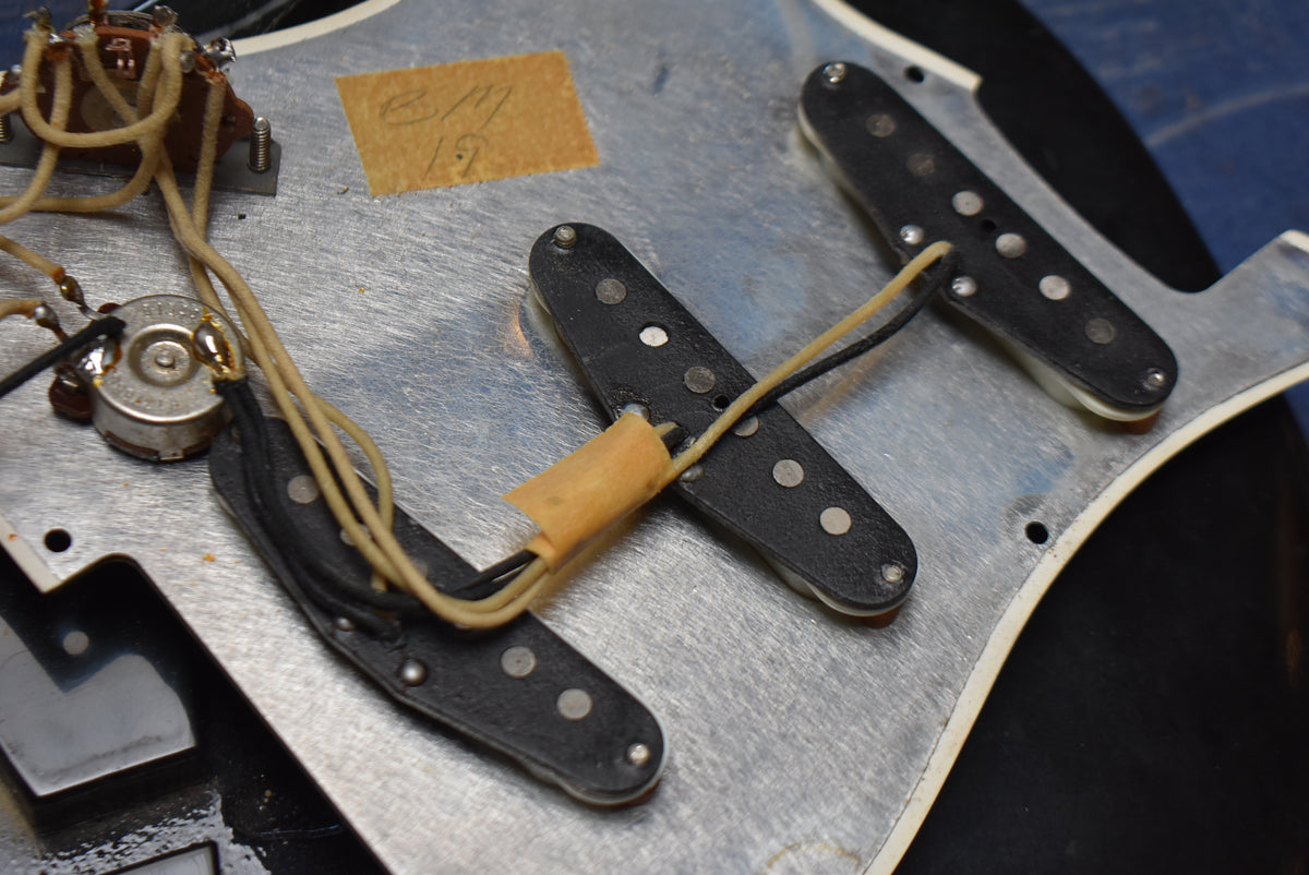 Close-up of the guitar's internal wiring, showing three black single-coil pickups, a capacitor, and control pots on a metal shielding plate.