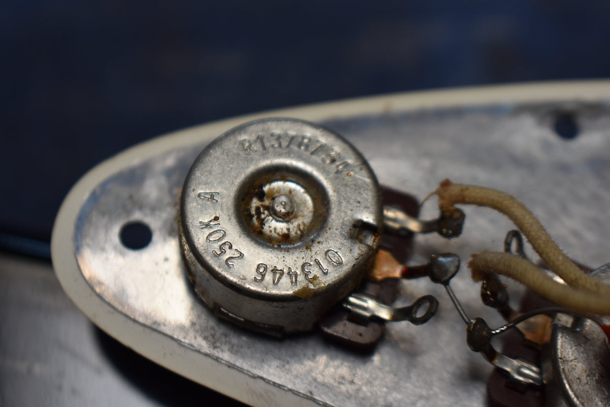 Close-up of a tarnished metal potentiometer with soldered wires, mounted on a worn white pickguard with a screw hole.