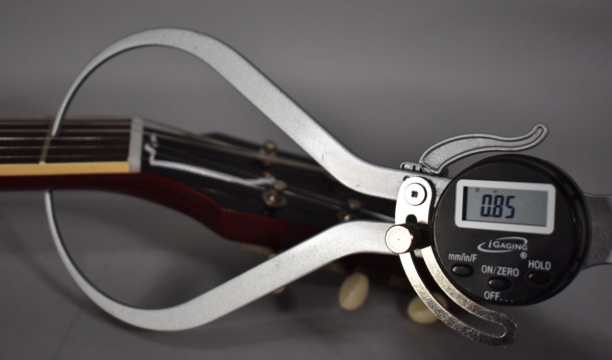 A digital caliper measures the string height at the first fret of the cherry red guitar, showing 0.85mm.