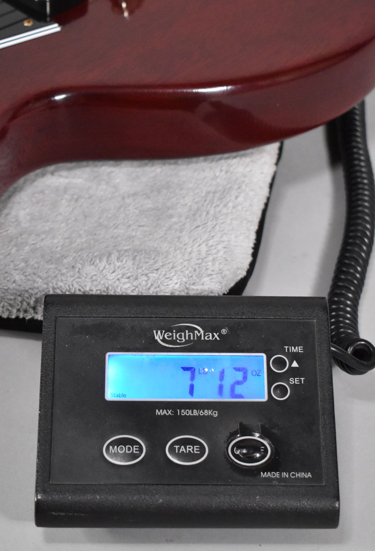 The cherry red guitar body rests on a towel, with a digital scale in the foreground displaying "7 LBS 12 OZ".