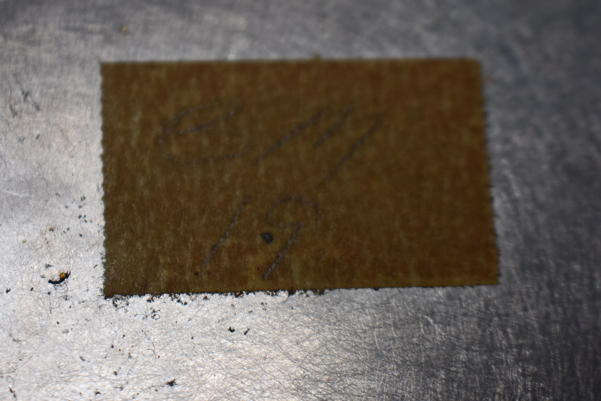 A rectangular piece of brown paper with faint pencil markings "88" and "19" on a scratched metal surface.
