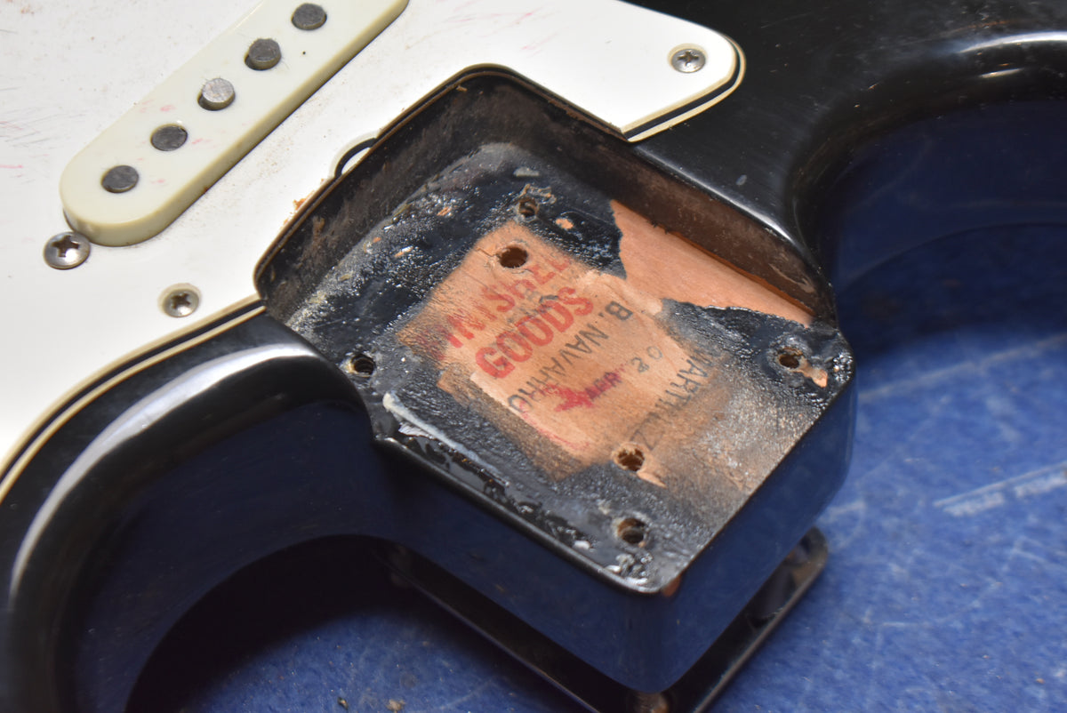 Close-up of the black guitar body's neck pocket, revealing a worn wooden interior with a faded red label and visible screw holes.