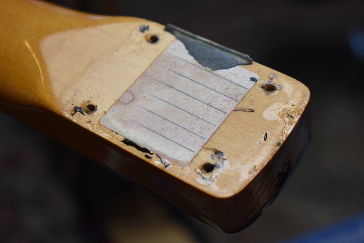 The heel of a light-colored guitar neck, showing four screw holes, a worn paper label, and black paint chipping from the body.