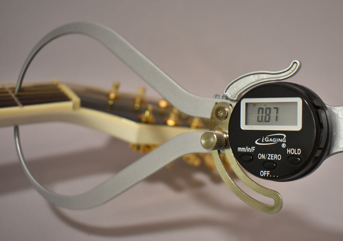 The guitar's pearl white headstock and gold tuners are visible, with a digital caliper measuring the nut width at 0.87.