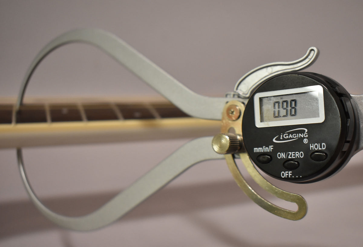 A digital caliper measures the neck thickness at the first fret, displaying "0.98" on its screen.