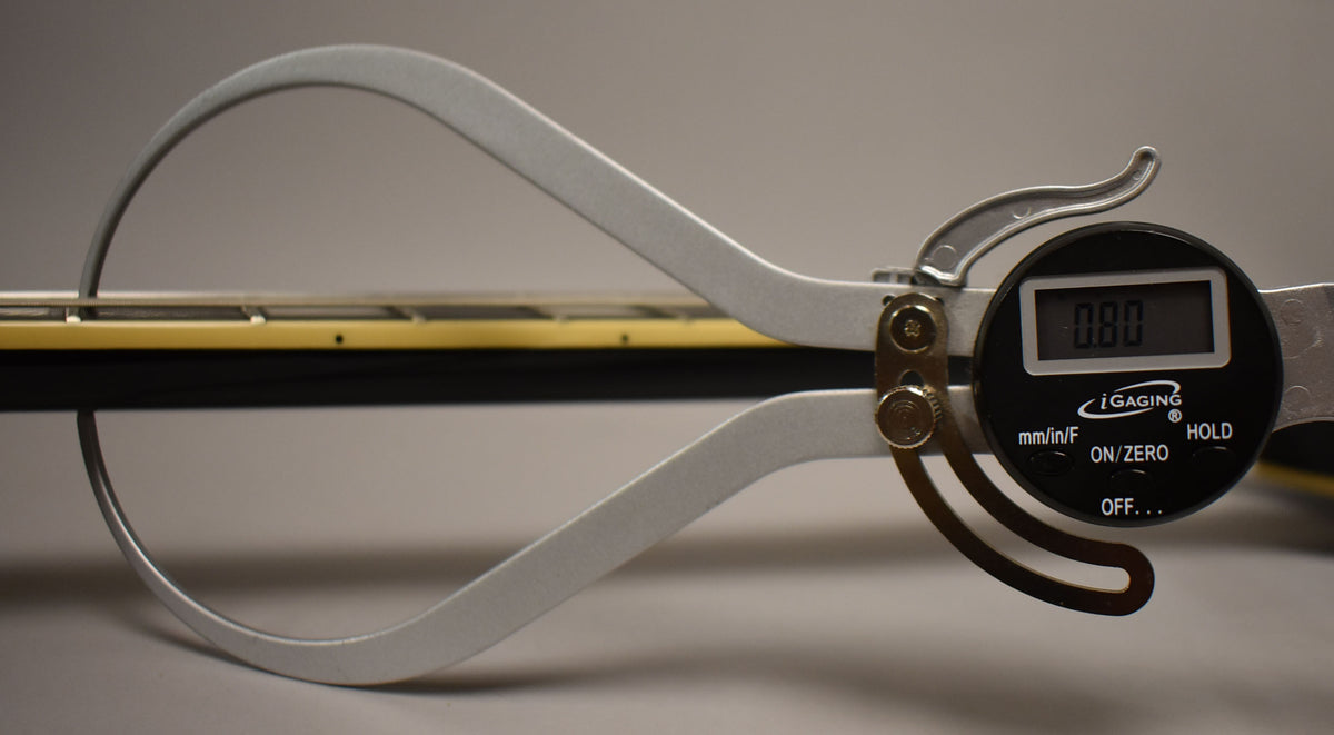 A digital caliper measures the guitar's neck, showing 0.80 on its screen, with the fretboard and strings visible.