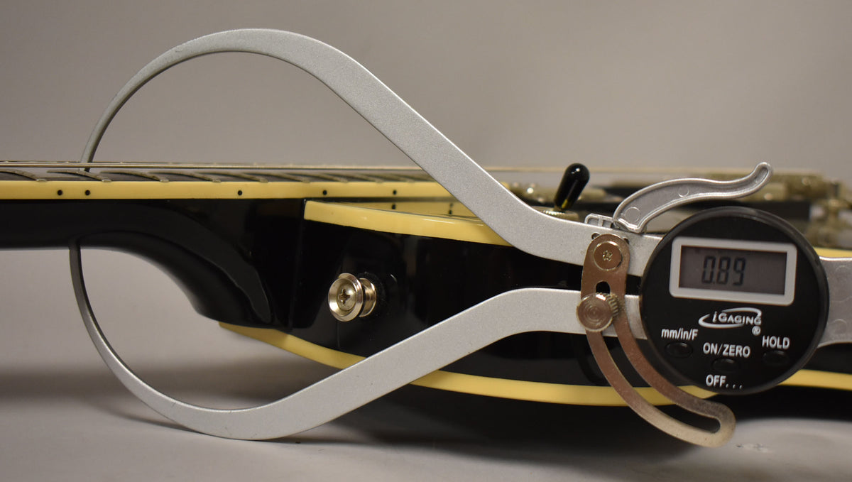 A digital caliper measures the string height at the 12th fret of the black guitar body with cream binding.