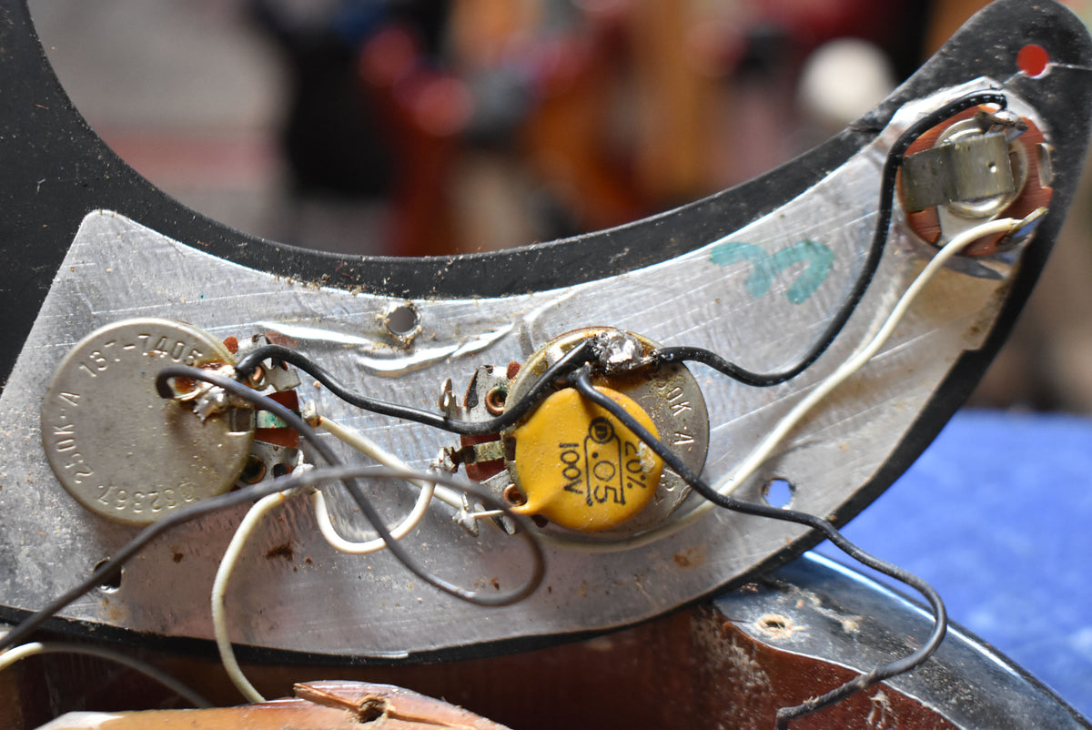 Close-up of a guitar's internal wiring, showing two potentiometers, a yellow capacitor labeled "0.05 100V 20%", and soldered connections.