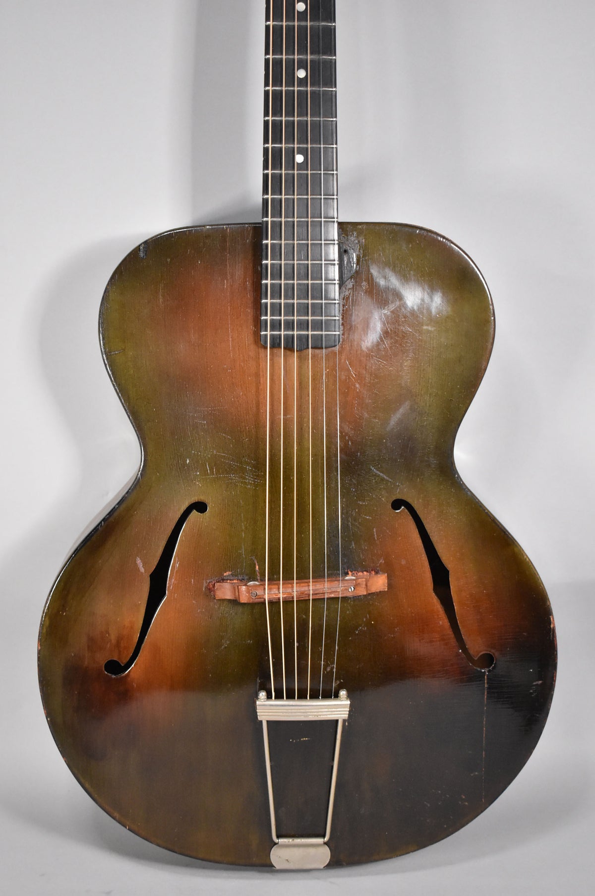 Full front view of the archtop guitar, showing its dark brown and green sunburst finish, f-holes, wooden bridge, and trapeze tailpiece.