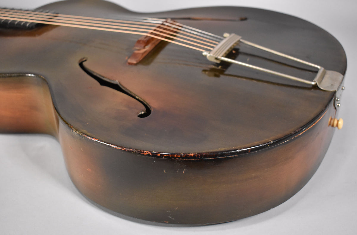 Close-up of the archtop guitar's lower bout, showing its dark brown sunburst finish, f-holes, and a metal trapeze tailpiece.