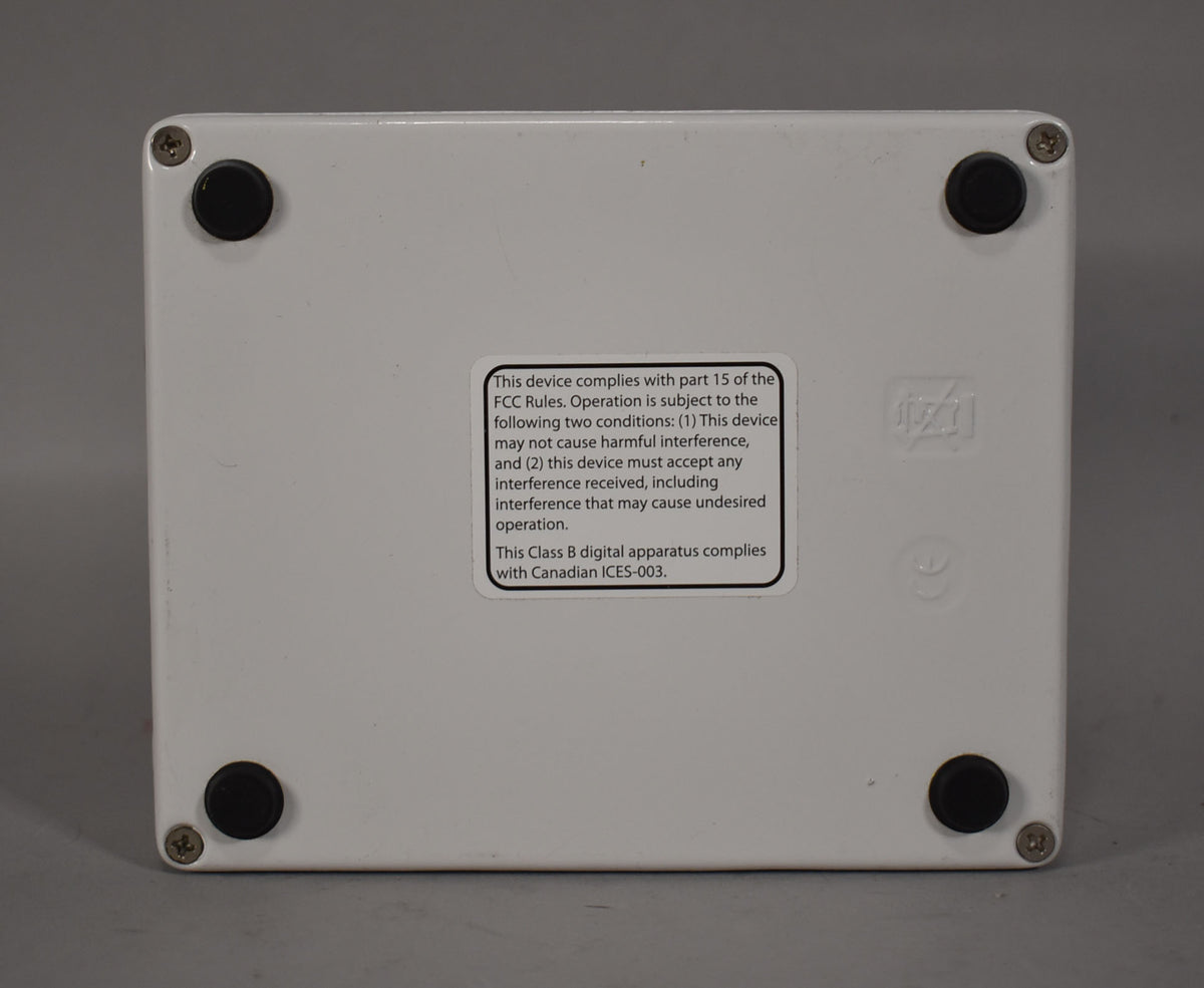 The white pedal's bottom is shown, featuring four black rubber feet, a compliance sticker, and faint scuffs.