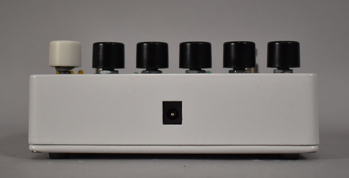 The white pedal is viewed from the rear, showing a power input jack and five control knobs, one cream and four black.