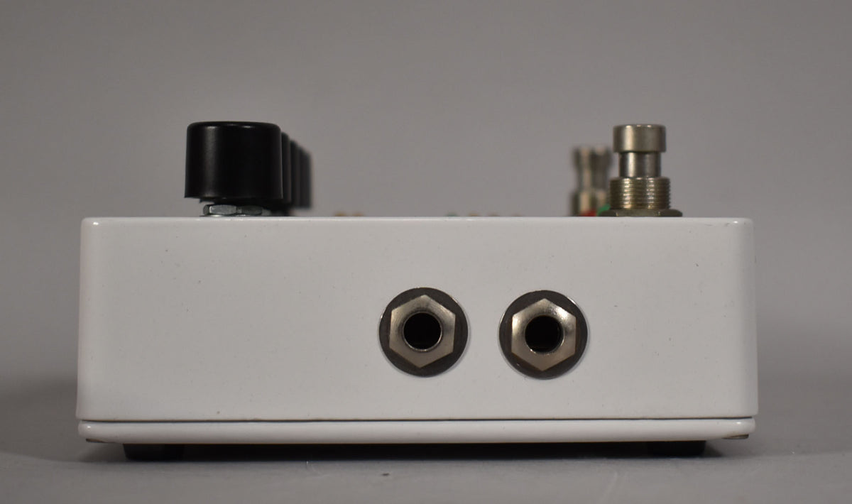 The white pedal is viewed from the side, showing two silver input jacks, a black knob, and a silver footswitch.