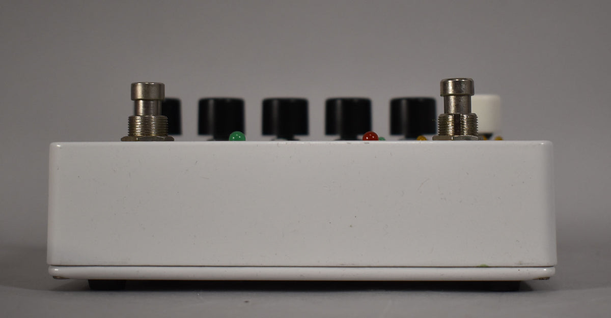 The white pedal is viewed from the side, revealing two silver footswitches, four black knobs, and two small green and red indicator lights.