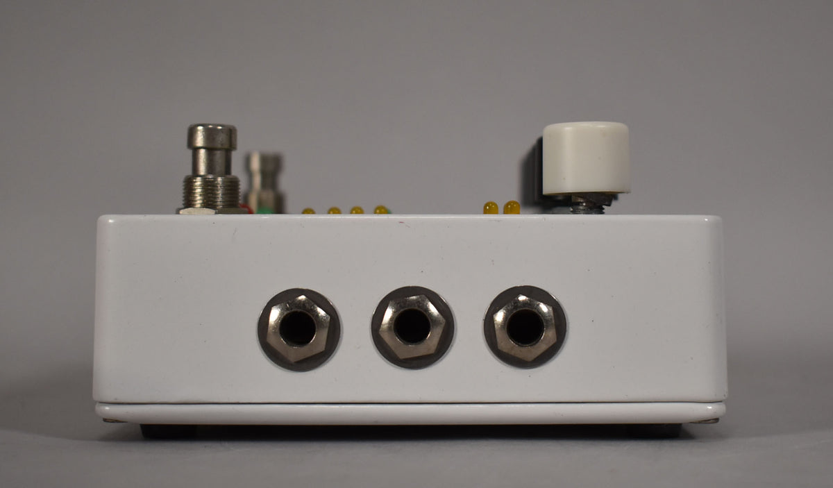 The white pedal is viewed from the side, showing three input jacks, a footswitch, a cream knob, and a row of amber LEDs.