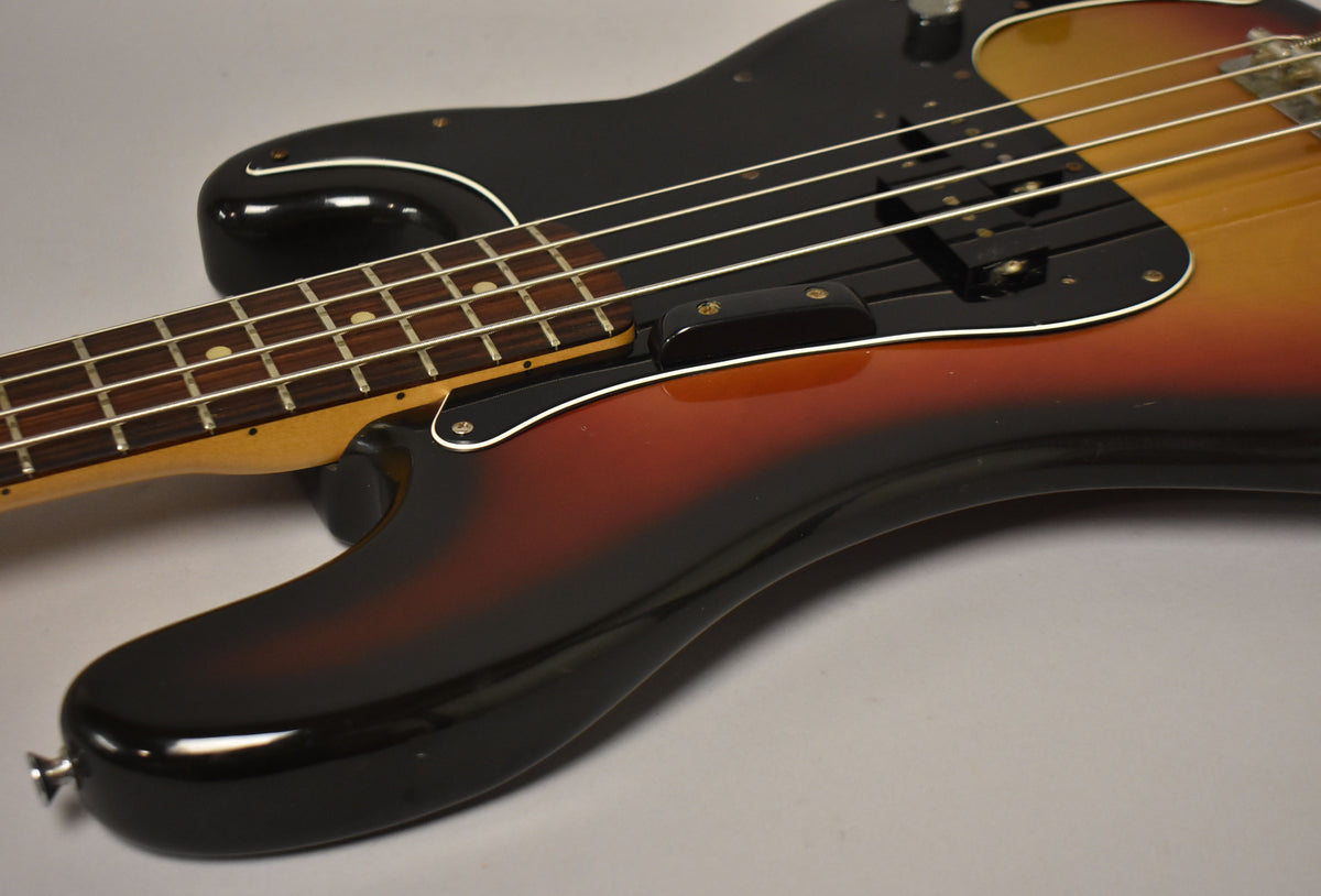 Close-up of a sunburst bass guitar body with a black pickguard, showing the lower bout, bridge, and part of the fretboard.