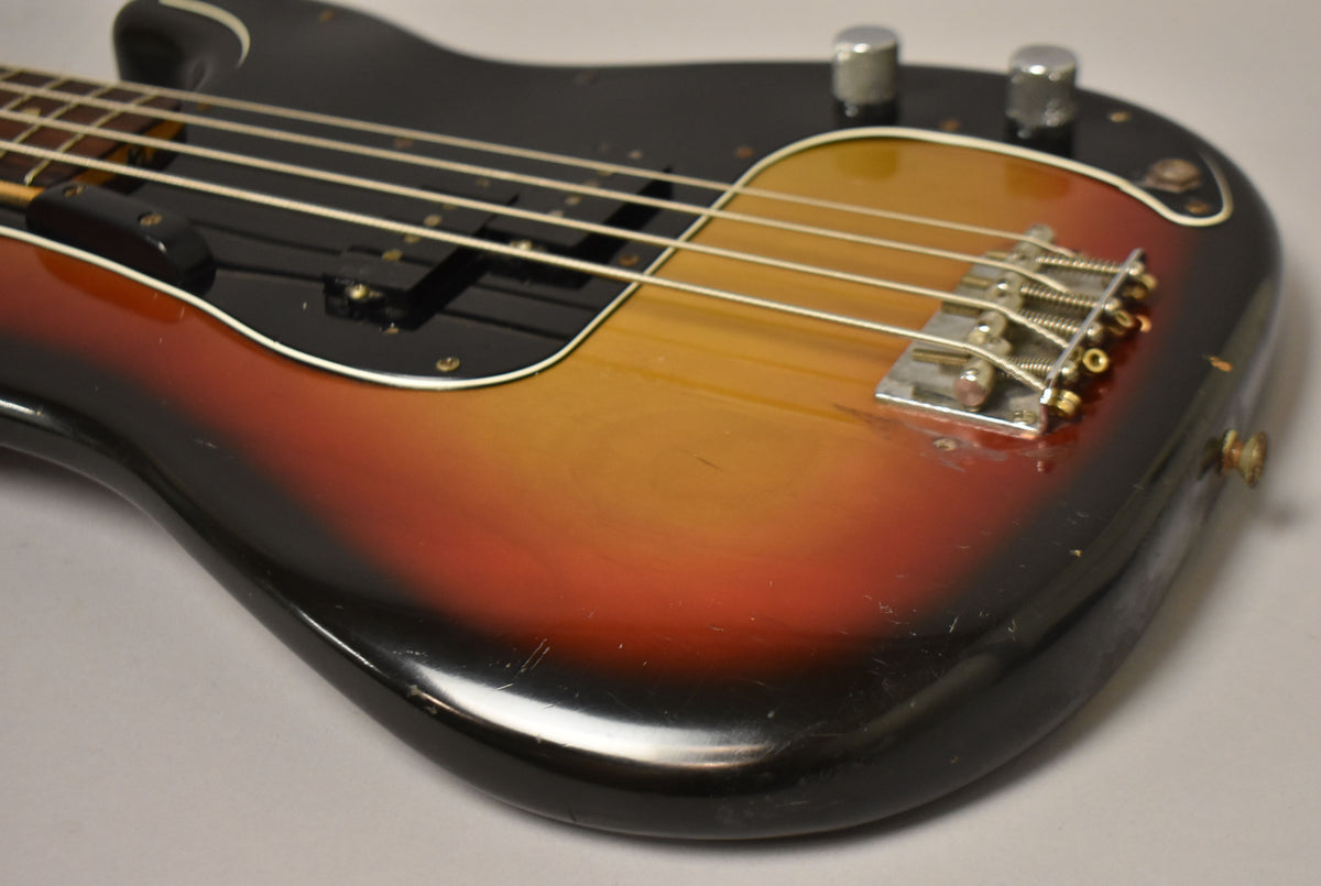 Close-up of a sunburst bass body with a black pickguard, showing a chrome bridge, two control knobs, and visible scratches and wear.