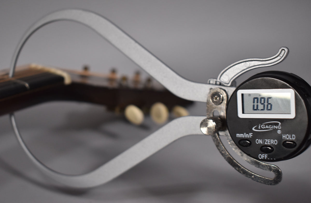 A digital caliper measures the dark brown neck and fretboard, showing a reading of 0.96, with tuning pegs blurred in the background.