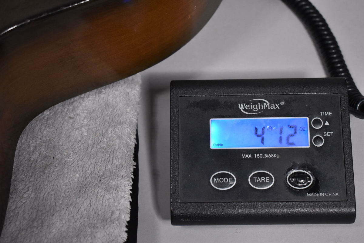The guitar's dark brown body rests on a gray towel, with a digital scale showing "4 LB 12 OZ" to the right.
