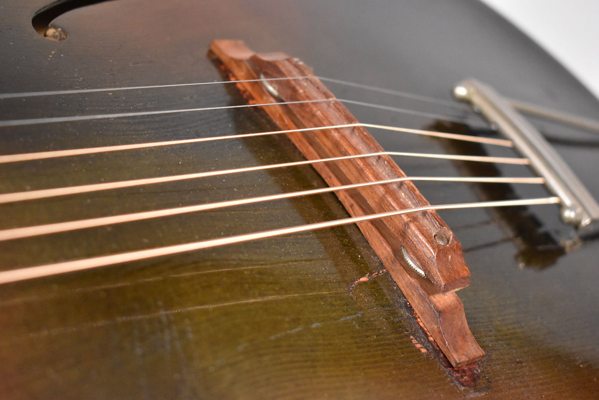 Close-up of a dark archtop guitar's wooden bridge, four strings, and part of a metal tailpiece, with wear on the body.