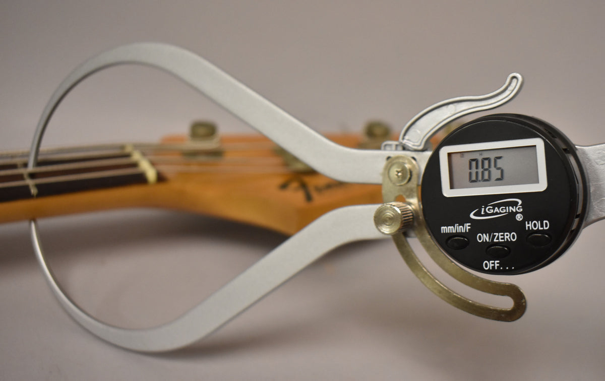 A digital caliper measures the bass's neck, showing "0.85" on its display, with the headstock blurred in the background.