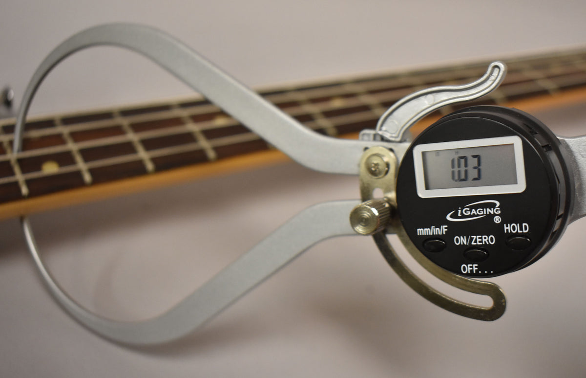 A digital caliper measures the bass's dark rosewood fretboard, showing "1.03" on its screen.