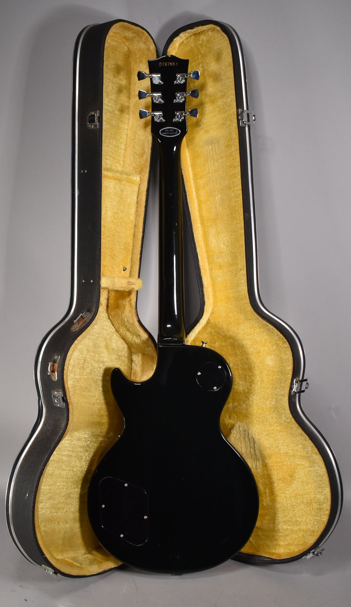 The black guitar is seen from the back, standing upright in its open, yellow-lined hard case, showing some light wear.