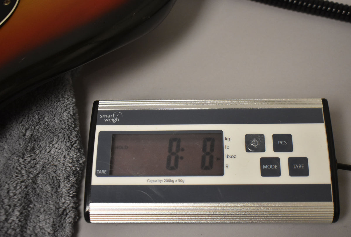 The sunburst bass body rests on a gray towel next to a digital scale displaying "8.8" pounds.