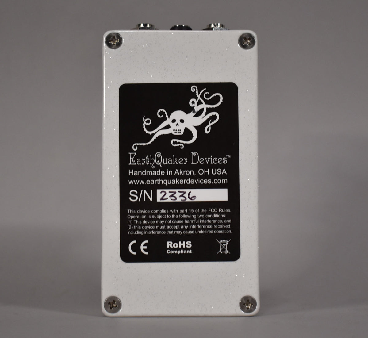 The pedal's white glitter-finish back panel is visible, with a black label featuring an octopus skull logo and serial number.