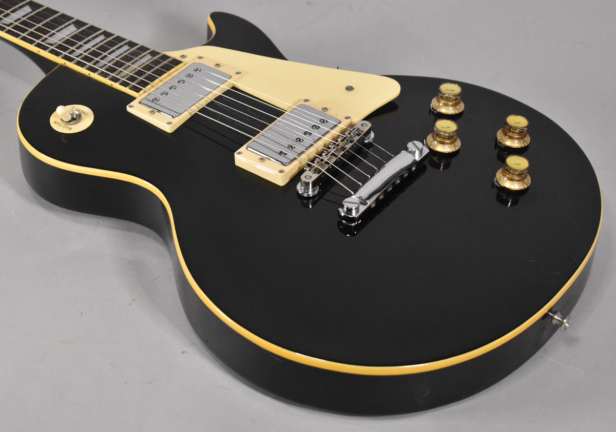 The black Les Paul style guitar is angled from the upper left, showing its cream binding, pickguard, and gold top hat knobs.