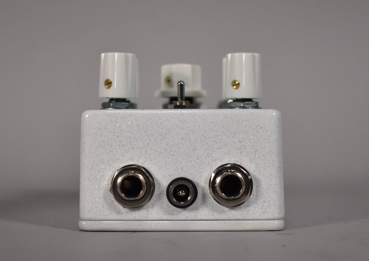 The white glitter-finish pedal is viewed from the front, showing three white knobs, a toggle switch, and three input jacks.