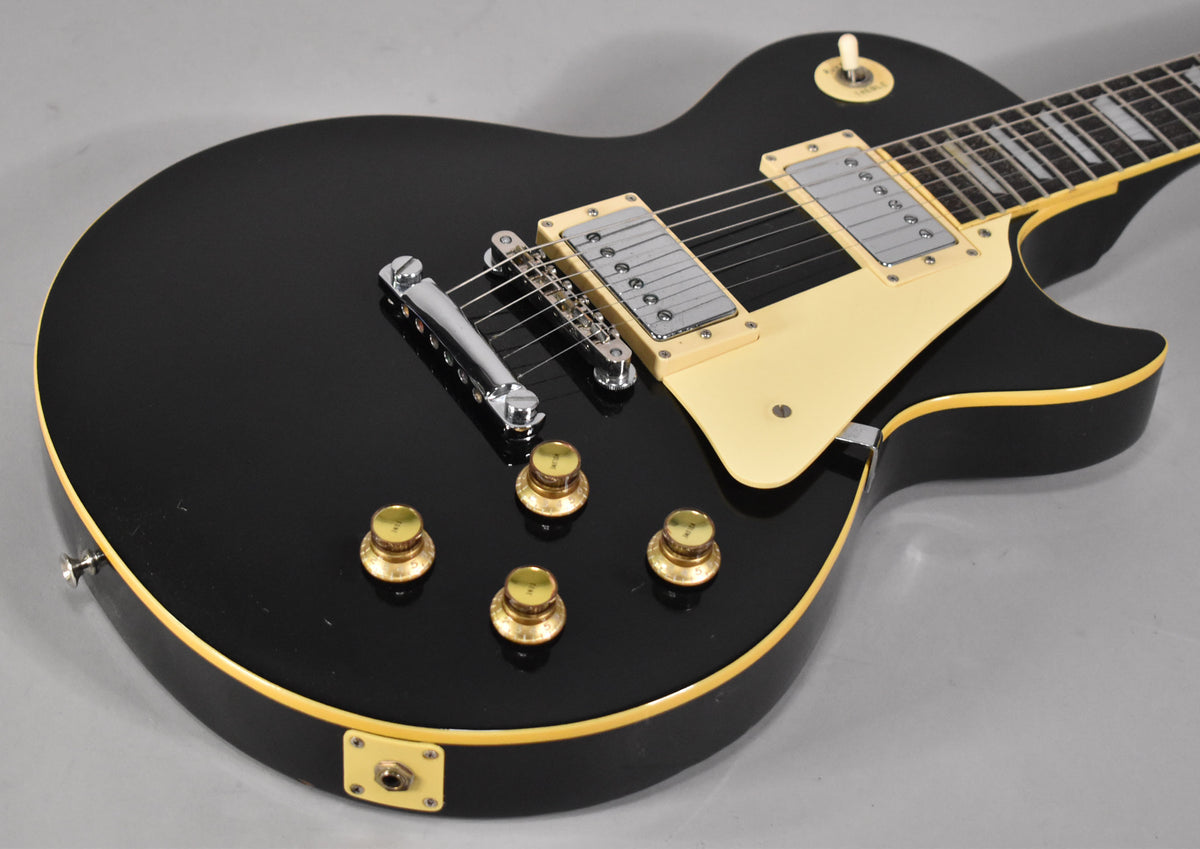 The black Les Paul copy is angled from the lower bout, showing its cream pickguard, two humbuckers, four gold speed knobs, and output jack.