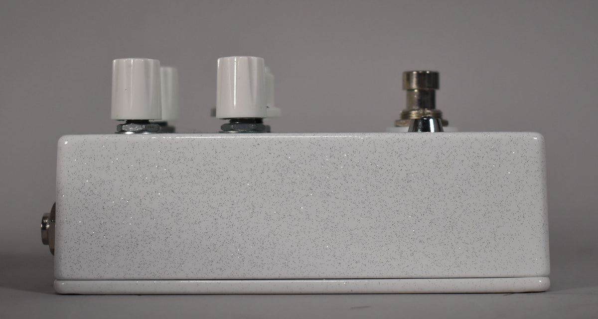 Side view of a white glitter-flecked pedal with three white knobs and a silver footswitch on top, and a silver jack on the left side.