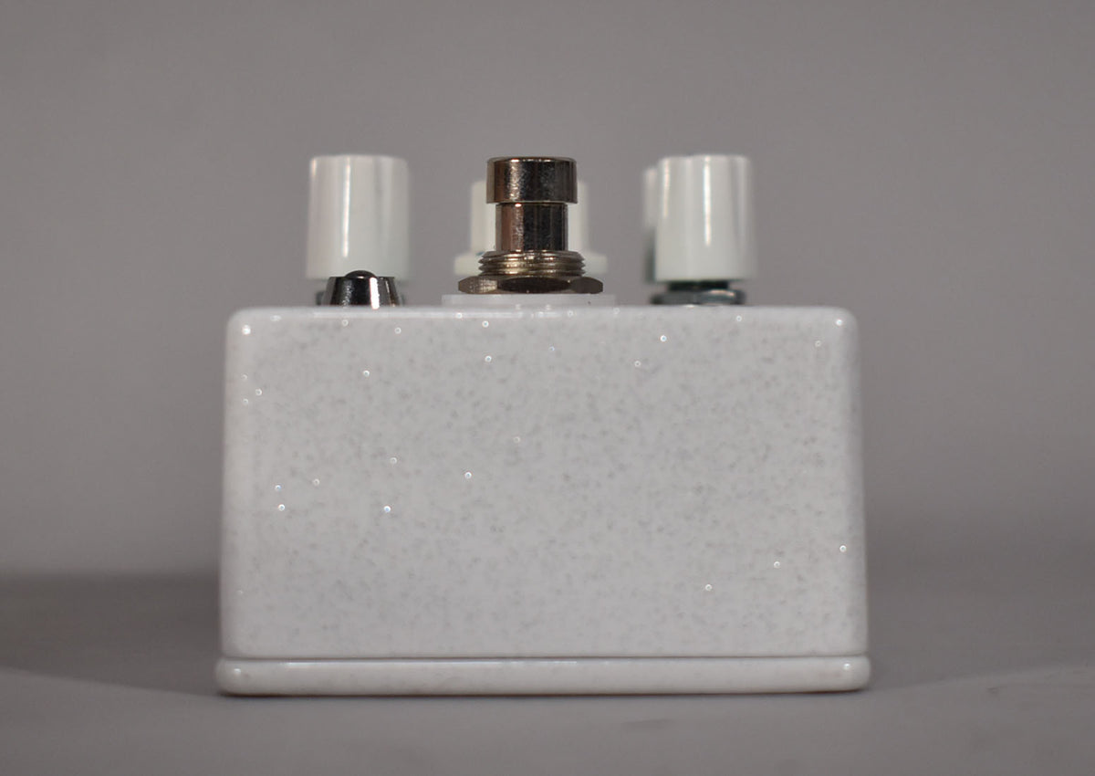 The white glitter-finish pedal is viewed from the front, showing three white knobs and a chrome footswitch.