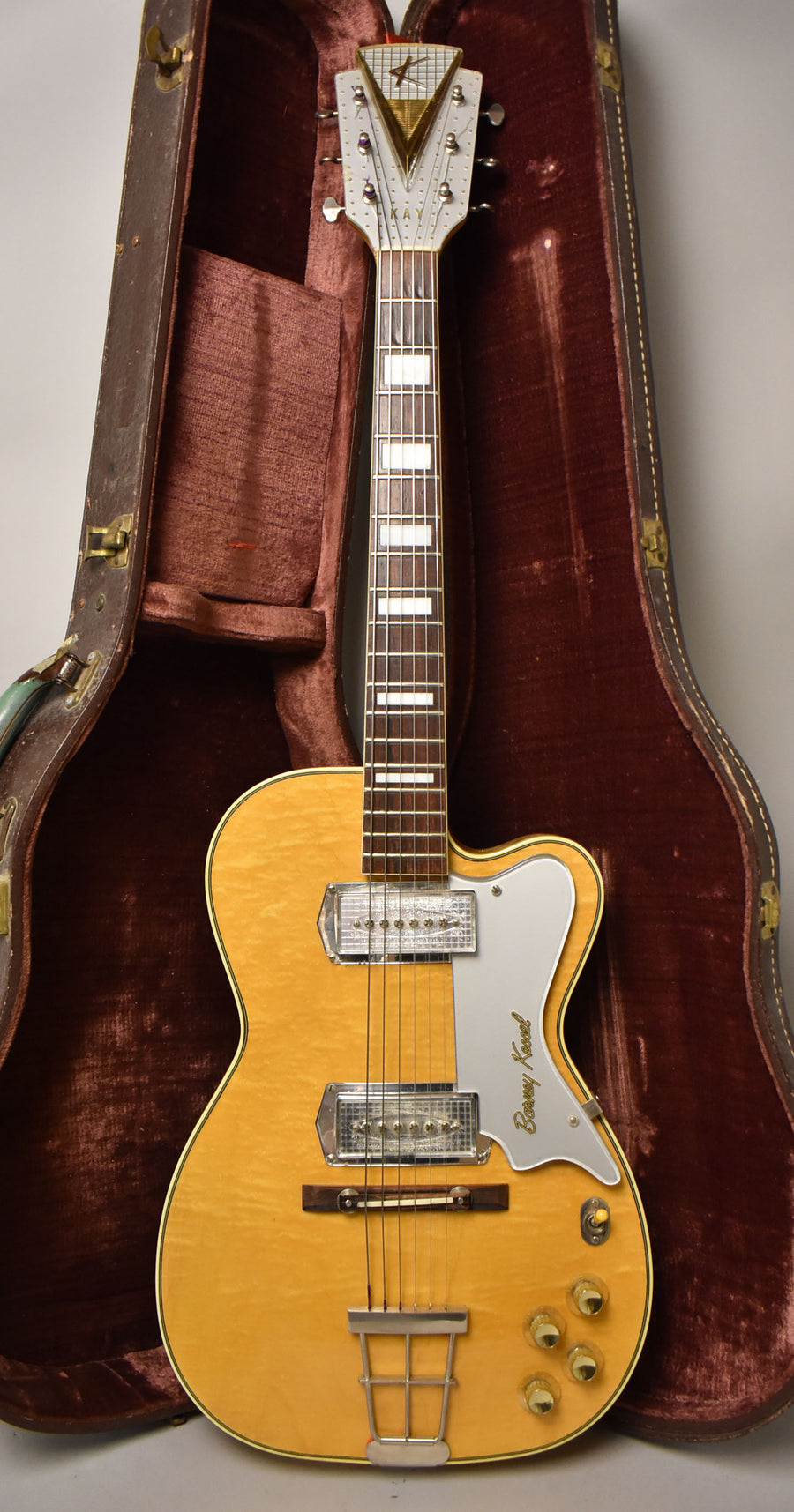 Full-length view of a blonde hollow-body electric guitar with a light pickguard, two clear pickups, and four gold knobs, in its open brown case.