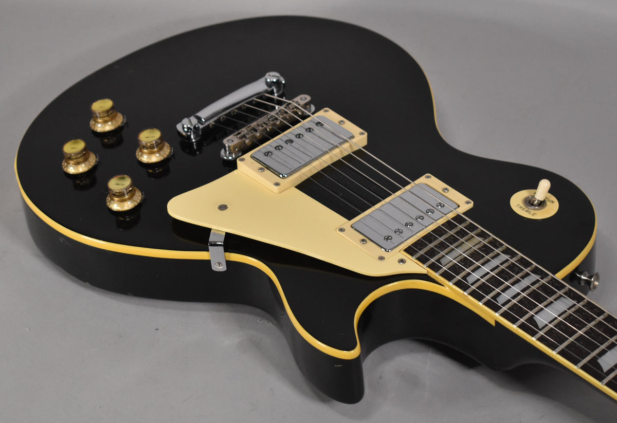 Black electric guitar body with cream binding, pickguard, pickup rings, and switch tip, showing gold knobs and chrome hardware.