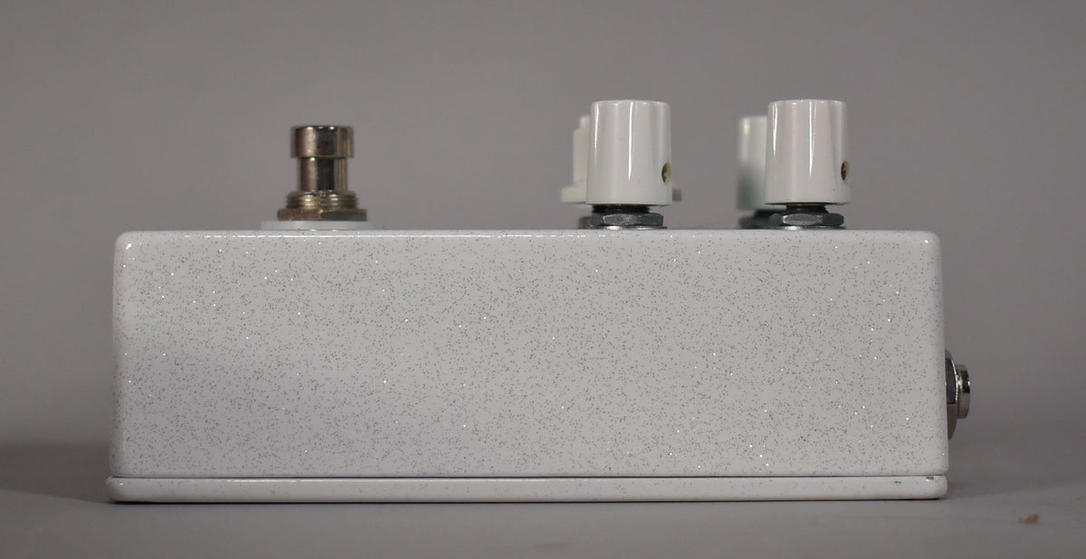 The white glitter-finish pedal is viewed from the right side, showing a footswitch, three white knobs, and an input jack.